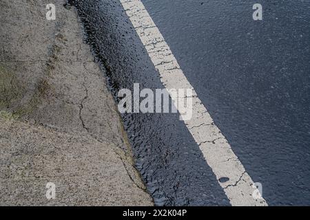 Nahaufnahme einer mit Wasser gefüllten Bordsteinkante entlang eines gerissenen Bürgersteigs und einer nassen Straße, die mit einer weißen Linie markiert ist. Zeigt städtische Entwässerung und Straßeninstandhaltung Stockfoto