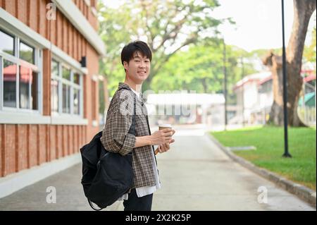 Ein junger asiatischer Student mit Rucksack steht vor einem Backsteinhaus auf dem Campus, hält eine Kaffeetasse und lächelt in die Kamera. Stockfoto