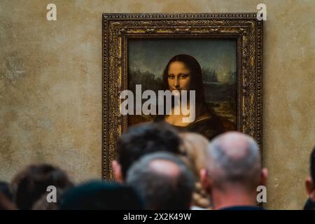 Eine Menge von Museumsbesuchern versammelte sich, um das ikonische Gemälde Mona Lisa zu sehen, das in einem kunstvollen goldenen Rahmen vor einer strukturierten Wand ausgestellt ist. Stockfoto