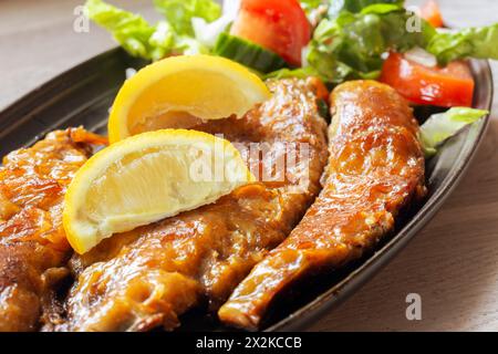 Geräucherte Makrelenfilets mit Salat und Zitrone auf einem schwarzen Teller. Auf einer Küchenarbeitsplatte bei spätabendlicher Sonneneinstrahlung Stockfoto