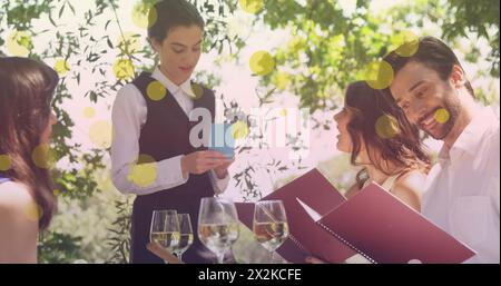 Bild von gelben Punkten über glücklichen kaukasischen weiblichen und männlichen Freunden, die im Restaurant bestellen Stockfoto