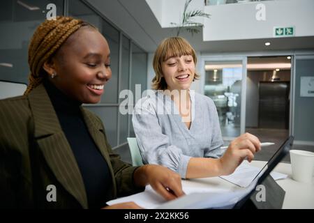 Zwei glückliche junge, multirassische Geschäftsfrau in Geschäftskleidung mit digitalem Tablet und Dokumenten im Büro Stockfoto