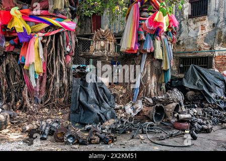 Gebrauchte Fahrzeugmotorteile sind um 2 Bodhi-Bäume verstreut, die mit einem bunten Tuch und einem Phra Phum-Schrein in der Mitte verbunden sind. Stockfoto