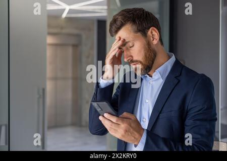 Ein besorgter Geschäftsmann in einem modernen Büro, der gestresst aussieht, während er eine Nachricht auf seinem Smartphone liest. Das Bild fängt einen Moment der Angst und der beruflichen Herausforderung ein. Stockfoto