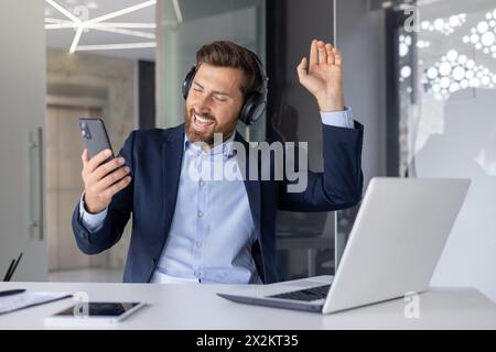 Fröhlicher Geschäftsmann mit Kopfhörern, der auf seinem Smartphone in einer modernen Büroumgebung zu Musik tanzt und singt. Freude und Entspannung am Arbeitsplatz ausdrücken. Stockfoto