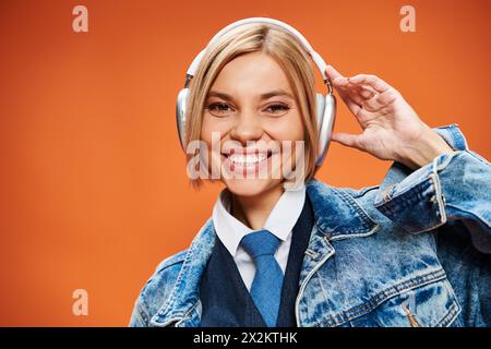 Fröhliche junge Frau mit blonden Haaren und Kopfhörern in Jeansjacke, die vor orangefarbenem Hintergrund posiert Stockfoto