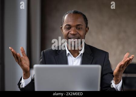 Ein reifer afrikanischer Geschäftsmann im Anzug spricht und Gesten vor einem Laptop während eines Online-Meetings. Er wirkt engagiert und proaktiv. Stockfoto