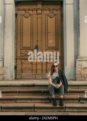 Junge Frau, die auf einer Treppe des Konzertsaals „Ave Sol“ (früher St. Peter und St. Paul orthodoxe Kirche) sitzt, mit Holztür auf einem Hintergrund 28.03.20 Stockfoto