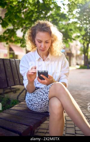 Schöne Frau mittleren Alters in 70er, 80er Jahre Stil Kleidung schaut auf ihr Smartphone auf einer Bank Stockfoto