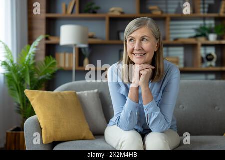Eine reife Frau genießt einen friedlichen Moment zu Hause. Ihr lächelndes Gesicht, das bequem auf einem Sofa sitzt, drückt Zufriedenheit und Freude aus, umgeben von einem geschmackvoll eingerichteten Wohnzimmer. Stockfoto