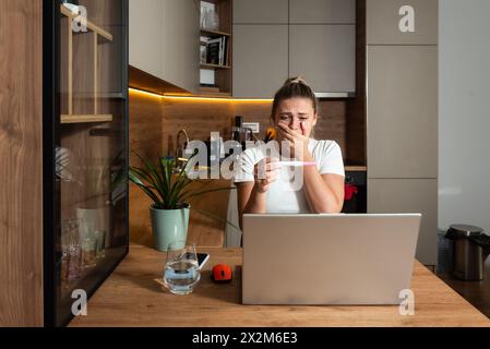 Enttäuschte junge frustrierte und schockierte schwangere Frau, die den Schwangerschaftstest in der Hand verwirrt und depressiv hält und die Mutter per Videoanruf auf dem Laptop anruft Stockfoto