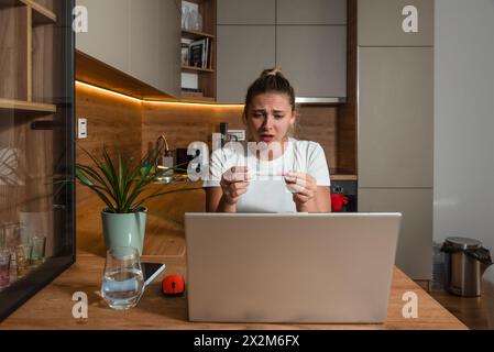 Enttäuschte junge frustrierte und schockierte schwangere Frau, die den Schwangerschaftstest in der Hand verwirrt und depressiv hält und die Mutter per Videoanruf auf dem Laptop anruft Stockfoto