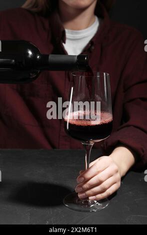 Alkoholsucht. Frau, die Rotwein aus der Flasche ins Glas gießt, an einem dunklen, strukturierten Tisch, Nahaufnahme Stockfoto