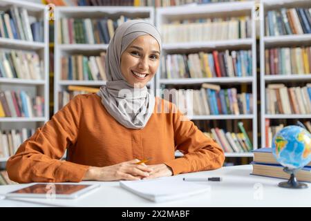 Eine fröhliche muslimische Frau im Hijab sitzt an einem Tisch in einer Bibliothek, umgeben von Regalen voller Bücher. Ihr Ausdruck vermittelt Selbstvertrauen und Glück, während sie sich an ihrem Studium beteiligt. Stockfoto