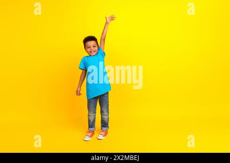 Foto in voller Größe des netten kleinen Schuljungen mit Afro-Haarbekleidung blaues stilvolles T-Shirt, das die Hand hoch hebt isoliert auf lebhaftem gelbem Hintergrund Stockfoto