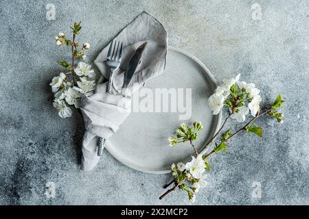 Blick von oben auf einen stilvollen Tisch mit Kirschblüten, Keramikplatte, silbernem Besteck und einer Leinenserviette auf strukturierter Oberfläche Stockfoto