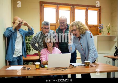 Eine Gruppe fröhlicher Senioren versammelt sich um einen Laptop mit animierten Ausdrücken und teilt ein Lernerlebnis in einer gemütlichen Umgebung im Klassenzimmer Stockfoto