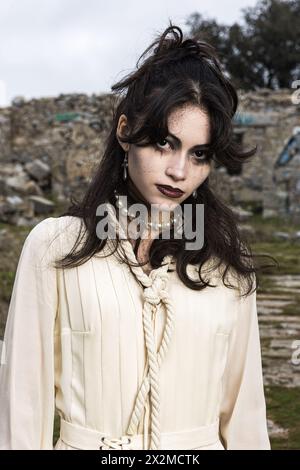 Eine junge Frau mit Gothic-inspiriertem Make-up steht vor einer Steinstruktur und trägt ein Vintage-Creme-Outfit mit Perlenakzenten Stockfoto