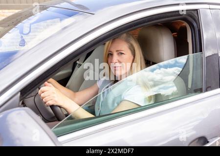 Eine selbstbewusste, reife Frau, die hinter dem Steuer eines Autos sitzt, in die Kamera lächelt, bereit für eine Reise Stockfoto
