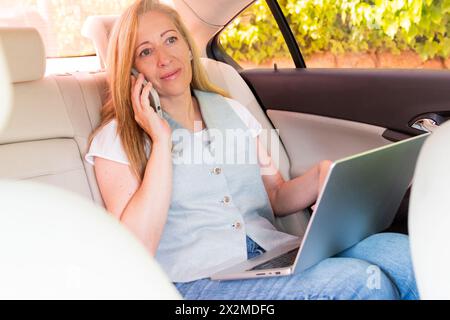 Eine erwachsene Frau, die auf dem Rücksitz eines Autos sitzt und während der Arbeit an einem Laptop telefoniert Stockfoto