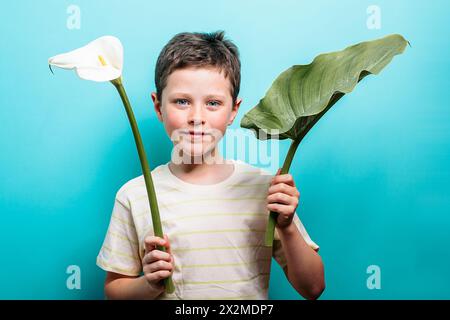 Ein fröhlicher kleiner Junge mit einem dezenten Lächeln steht vor einem leuchtend blauen Hintergrund, der eine weiße Calla-Lilie und ein großes grünes Blatt hält Stockfoto