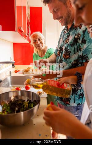 Eine fröhliche ältere Frau sieht zu, wie ein Mann Olivenöl in eine Salatschüssel gießt, mit Zutaten um sie herum, und eine andere Frau hilft, Essen für ein f zuzubereiten Stockfoto