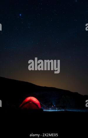 Ein beleuchtetes Zelt unter dem Sternenhimmel schafft eine ruhige Campingszene in Fuerteventura, Spanien, mit einer Kulisse aus silbrigen Bergen. Stockfoto
