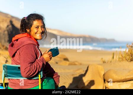 Eine freudige Frau, die sich mit einer Tasse in der Hand beim Camping an den Sandstränden von Fuerteventura, den Kanarischen Inseln, entspannt. Stockfoto