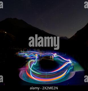 Eine Nachtszene, die farbenfrohe Lichtwege auf einem gewundenen Radweg festlegt, mit Bergen und Sternenhimmel im Hintergrund, während der Sommer Stockfoto