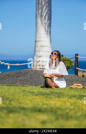 Eine Frau genießt einen friedlichen Moment in einer natürlichen Umgebung, liest ein Buch, während sie auf dem Gras sitzt, mit einem klaren blauen Himmel über dem Kopf und dem Meer im B Stockfoto