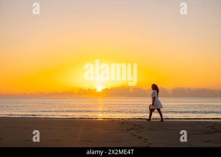 Eine Frau genießt einen ruhigen Spaziergang entlang eines Sandstrandes, während die Sonne aufgeht und ein Gefühl der Ruhe und Verbindung mit der Natur reflektiert. Stockfoto