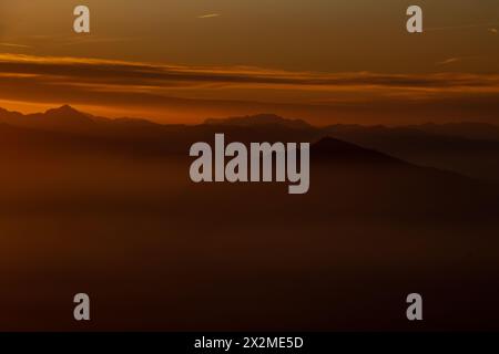 Ein ruhiger, nebeliger Blick auf die Silhouette der Gipfel rund um den Lago Maggiore, der in warmen Sonnenaufgangstönen getaucht ist Stockfoto