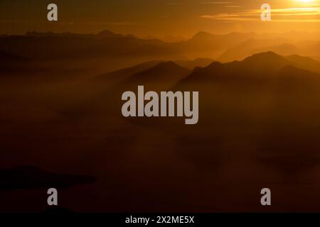 Ein ruhiger Blick fängt die goldenen Sonnenaufgangsstrahlen ein, die durch den Nebel über die bergige Landschaft des Lago Maggiore dringen Stockfoto