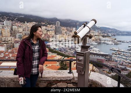 Eine junge Frau genießt mit einem Münzteleskop im Vordergrund die malerische Aussicht auf Monacos Stadtlandschaft und den Hafen Stockfoto