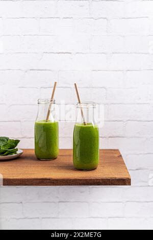 Zwei nährstoffreiche grüne Smoothies in Klarglasflaschen mit Papierhalmen auf einem Holzregal, an einer weißen Ziegelwand Stockfoto