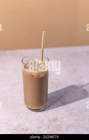Ein hohes Glas cremigen Eiskaffee, komplett mit Eiswürfeln und einem umweltfreundlichen Stroh, auf einer Marmoroberfläche mit einem warmen beigefarbenen Hintergrund Stockfoto