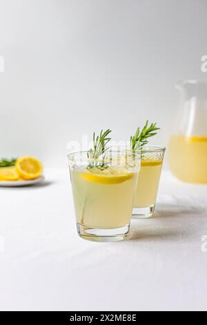 Zwei Gläser frisch zubereitete Limonade mit Rosmarinzweigen und einer geschnittenen Zitrone im Hintergrund auf einer sauberen weißen Oberfläche Stockfoto