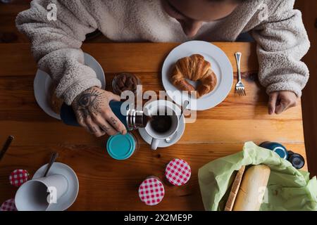 Die Person genießt ein warmes Frühstück mit Croissants, Kaffee und Mandelmilch und zeigt eine tätowierte Hand Stockfoto