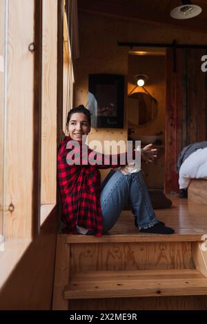 Eine fröhliche junge Frau in einem roten karierten Hemd und Jeans sitzt auf Holztreppen in einer rustikalen Hütte und lächelt zufrieden Stockfoto