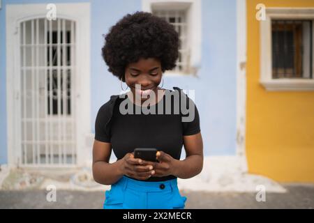 Eine fröhliche junge Afro-Frau, die ein schwarzes T-Shirt und eine blaue Hose trägt, benutzt ihr Handy mit einem Lächeln und steht auf einer Straße Stockfoto