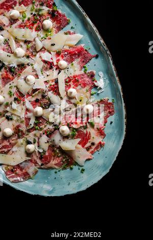 Von oben erlesenes Rindfleisch-Carpaccio mit geraspeltem Parmesan, garniert mit Kräuterstreuseln und dekorativen Speiseperlen auf einer Keramikplatte. Stockfoto