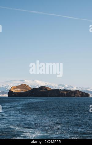 Ein ruhiger Blick auf die isländische Küste mit Klippen vor einem azurblauen Meer mit schneebedeckten Bergen im Hintergrund, unter einem klaren blauen Himmel Stockfoto