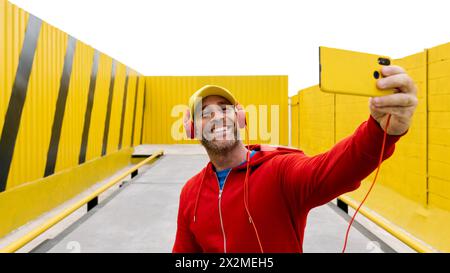 Ein fröhlicher Mann mit Mütze, Kopfhörern und roter Jacke macht ein Selfie mit hellgelbem Hintergrund Stockfoto