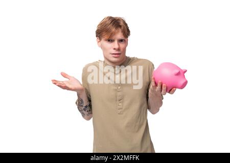 Junger rothaariger Mann mit Brille in braunem Hemd hält eine rosafarbene Schweinebank und entscheidet, wo er seine Ersparnisse ausgeben soll Stockfoto