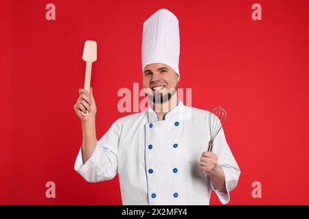 Glücklicher professioneller Konditor in Uniform mit Schneebesen und Spachtel auf rotem Hintergrund Stockfoto