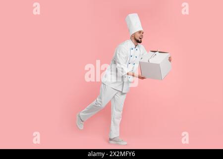 Glücklicher professioneller Konditor in Uniform mit Kuchenbox auf rosa Hintergrund Stockfoto