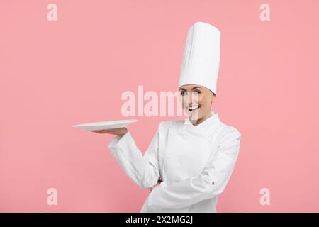 Glücklicher professioneller Konditor in Uniform, der leere Teller auf rosafarbenem Hintergrund hält Stockfoto