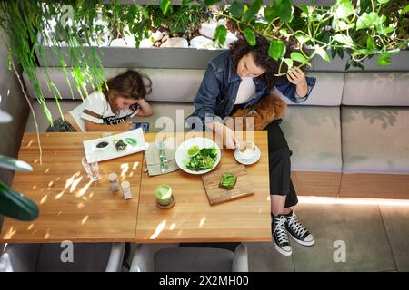 Familie Mutter Tochter und Hund, essen Sie im lebhaften veganen Restaurant, genießen Sie das Essen umgeben von üppigem Grün und genießen Sie pflanzlichen Lebensstil mit gesunder Ernährung Stockfoto