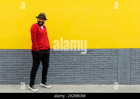 Erwachsener Latino-Mann (39) mit Bart, der mit den Händen in den Hosentaschen steht und eine fröhliche Geste macht. Er trägt einen Hut, einen roten Pullover und einen weißen Fl Stockfoto