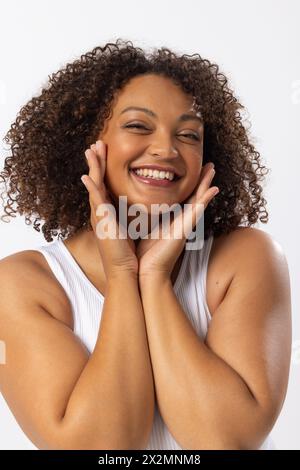 Birassisches Model in Übergröße mit lockigen braunen Haaren lächelt und berührt ihr Gesicht auf weiß Stockfoto
